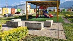 A playground with a slide and swings under a shelter, surrounded by a paved area with benches and greenery, with buildings and a mountain in the background. - Cozy 2-bedroom house in adorable Ahuachapan with AC / pool (Ahuachapan)