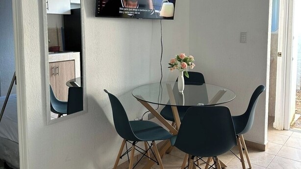 A dining area with a glass table and four chairs, a TV mounted on the wall, and a kitchen visible through an open door. - Cozy 2-bedroom house in adorable Ahuachapan with AC / pool (Ahuachapan)