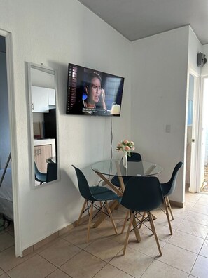 Un comedor con una mesa de vidrio y cuatro sillas, un televisor montado en la pared, y una cocina visible a través de una puerta abierta.