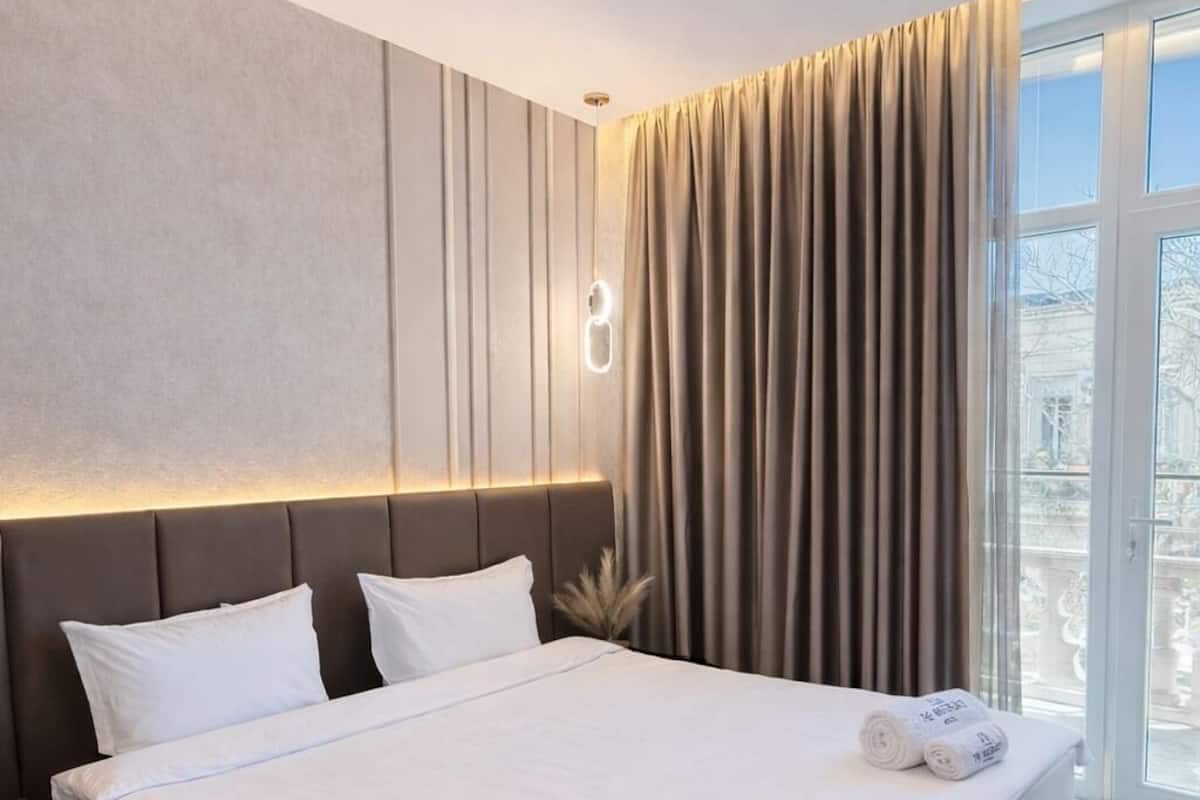 A modern hotel room with a large bed, a nightstand, a wall-mounted light, and a view of the outdoors through a large window.