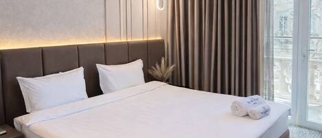 A modern hotel room with a large bed, a nightstand, a wall-mounted light, and a view of the outdoors through a large window.