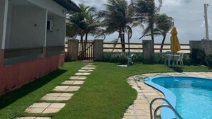 A swimming pool with a stone pathway leading to a covered patio area, surrounded by palm trees and a view of the beach. - POUSADA SOLEIL DE GENIPABU (Extremoz)