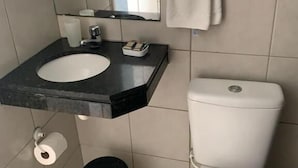A bathroom with a white toilet, a sink with a black countertop, a mirror, a towel rack with a towel, a trash bin, and a toilet paper holder. - POUSADA SOLEIL DE GENIPABU (Extremoz)