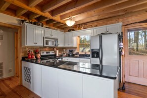 A kitchen with white cabinets, a black countertop, a stainless steel refrigerator, and a wooden ceiling. - Whitetail Bluff Cabin (Gilmer County)