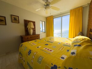 Ein Schlafzimmer mit Bett, Deckenventilator, Fenster mit gelben Vorhängen und Blick auf die Stadt.