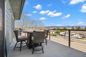 A balcony with wicker chairs and a table, overlooking a street with RVs and a white picket fence. - This Is It | Ocean Views, Walk to Park & Beach (Crescent City)