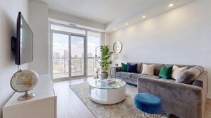 A modern living room with a large window, a grey sofa with multiple cushions, a glass coffee table, and a globe on a white cabinet. - Luxury 2-Bed London Apartment Greenwich & Canary Wharf (London)