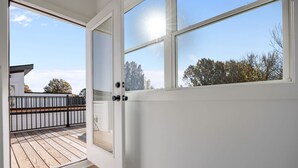 Puerta abierta que da a un balcón con terraza de madera y vistas a árboles y un edificio.