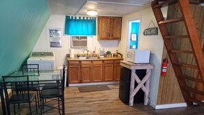 A compact kitchen with wooden cabinets, a built-in oven, and a microwave. There is a dining area with a glass table and chairs. A wooden ladder is visible in the corner. - Lakeside Cabin #4 (Eastland County)