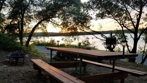 A picnic table with benches, a fire pit, and a grill set up by a lakeside. - Lakeside Cabin #4 (Eastland County)