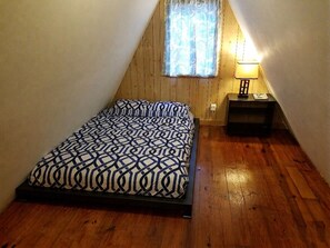 Una habitación pequeña con techo inclinado, una cama, una mesita de noche, una lámpara y un ventanal con cortinas.