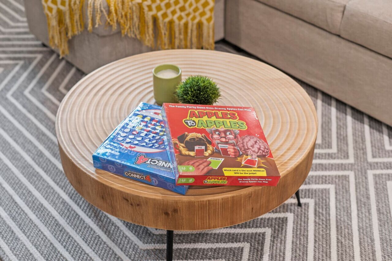 Een ronde houten salontafel met twee gezelschapsspellen, een kleine plant in pot en een groene mok.