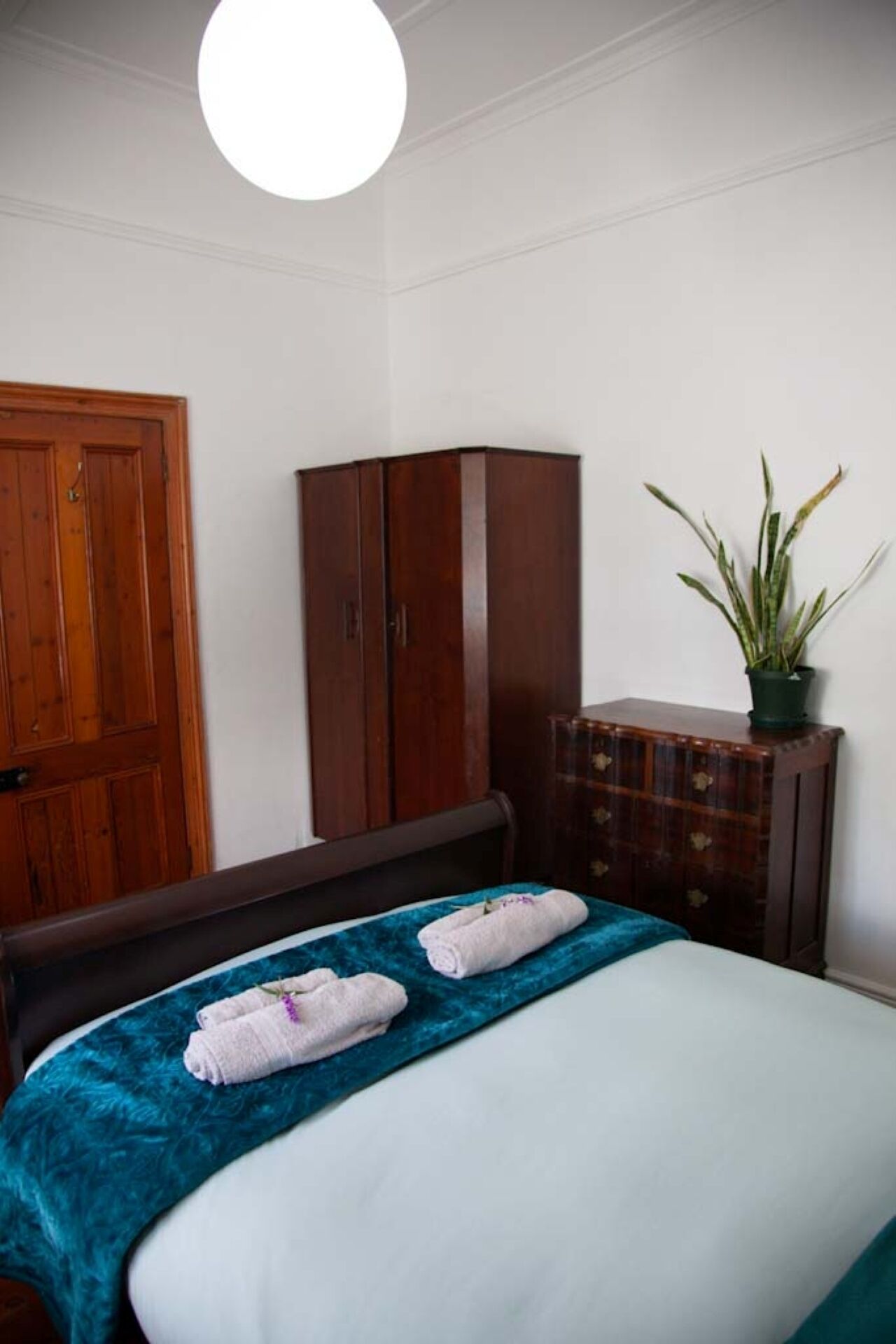 A bedroom with a bed, a wooden wardrobe, a cabinet, and a potted plant.