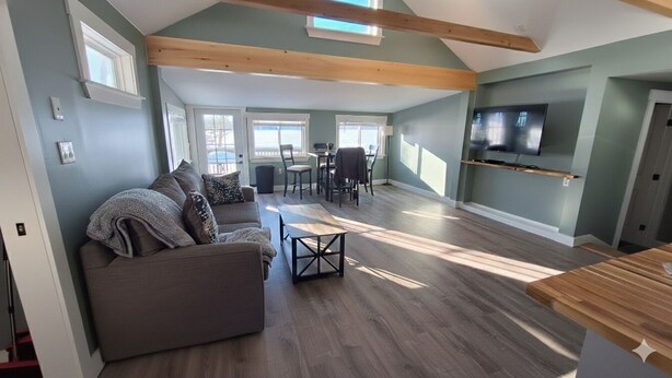 A spacious living area with a grey sofa, wooden beams, a dining table with chairs, and a TV mounted on the wall. - Modern Hermon Pond Retreat: 25ft to Shore, 15m to Bangor, Fishing & Kayak Launch (Hermon)