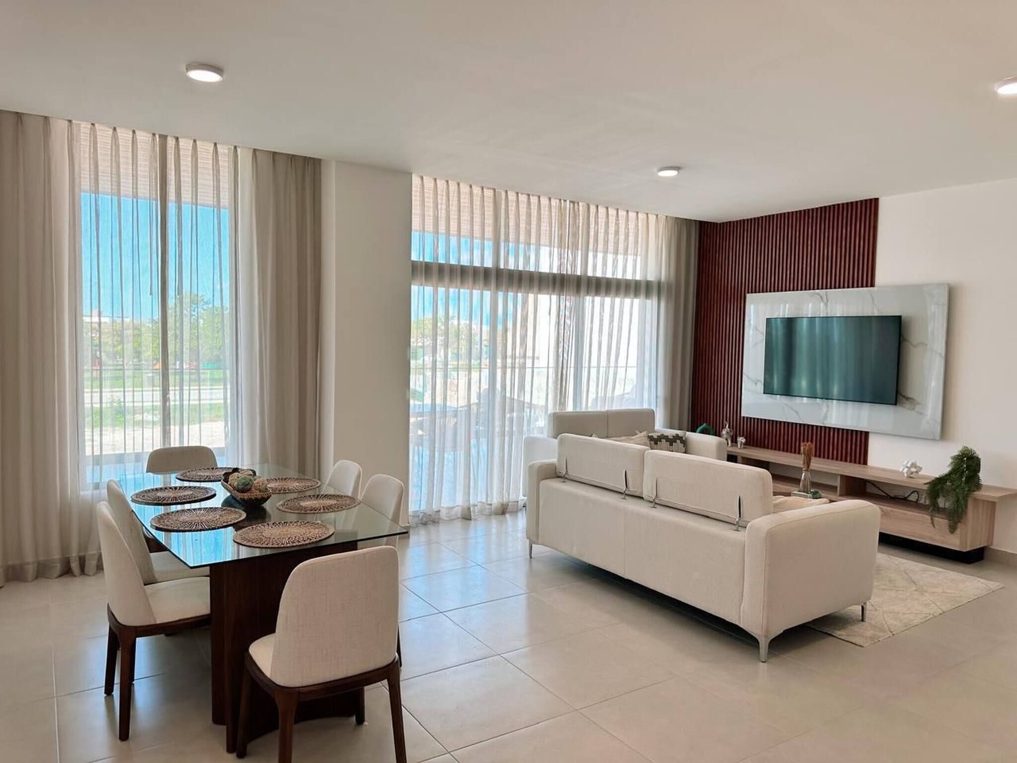 A modern living room with a glass dining table, white sofa, and a flat-screen TV mounted on the wall.