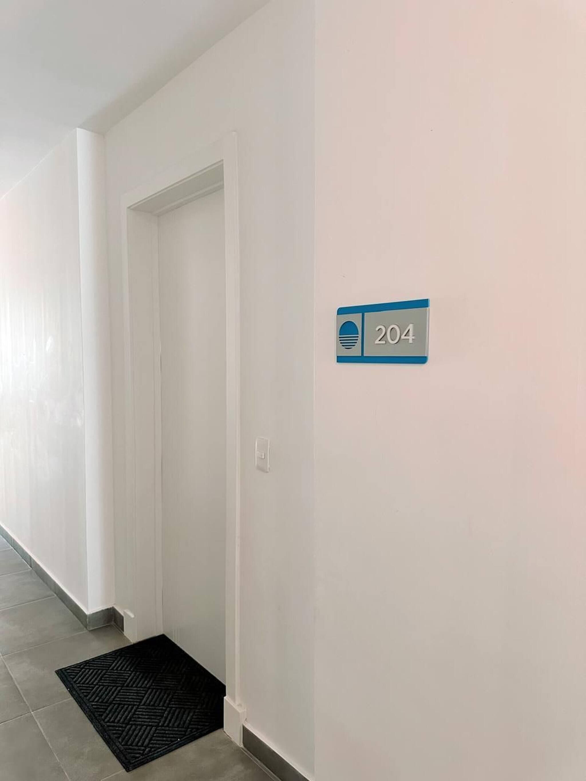 A hallway with a white door, a black doormat, and a blue sign with the number 204 on a white wall.