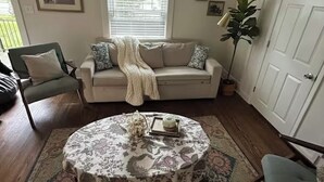 Sala de estar com sofá bege, mesa de centro redonda e tapete estampado. Há duas cadeiras, uma planta e uma porta ao fundo.