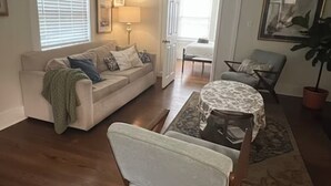 Sala de estar com sofá, uma cadeira, mesinha e planta em vaso. Há uma porta que dá para outro cômodo.