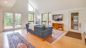 A spacious living room with a blue sofa, wooden flooring, large windows, and a television. - Peaceful Vermont Hideaway Near Stowe | Wooded View (Elmore)