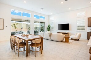 A modern living room with a dining area, a flat-screen TV, and a large window overlooking a garden. - Stylish Desert Home Mountain Views & Tesla Charger (Palm Springs)