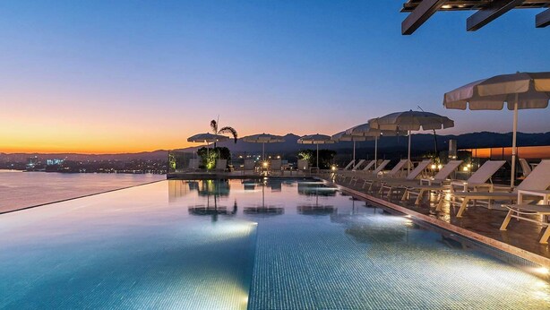 A swimming pool with lounge chairs and umbrellas, surrounded by a cityscape at sunset. - Panarea 5C | Beachfront 3BR + Panoramic Ocean View (Bucerías)