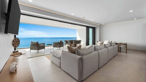 A modern living room with a large sofa, a flat-screen TV, and a dining area with a view of the ocean. - Panarea 5C | Beachfront 3BR + Panoramic Ocean View (Bucerías)