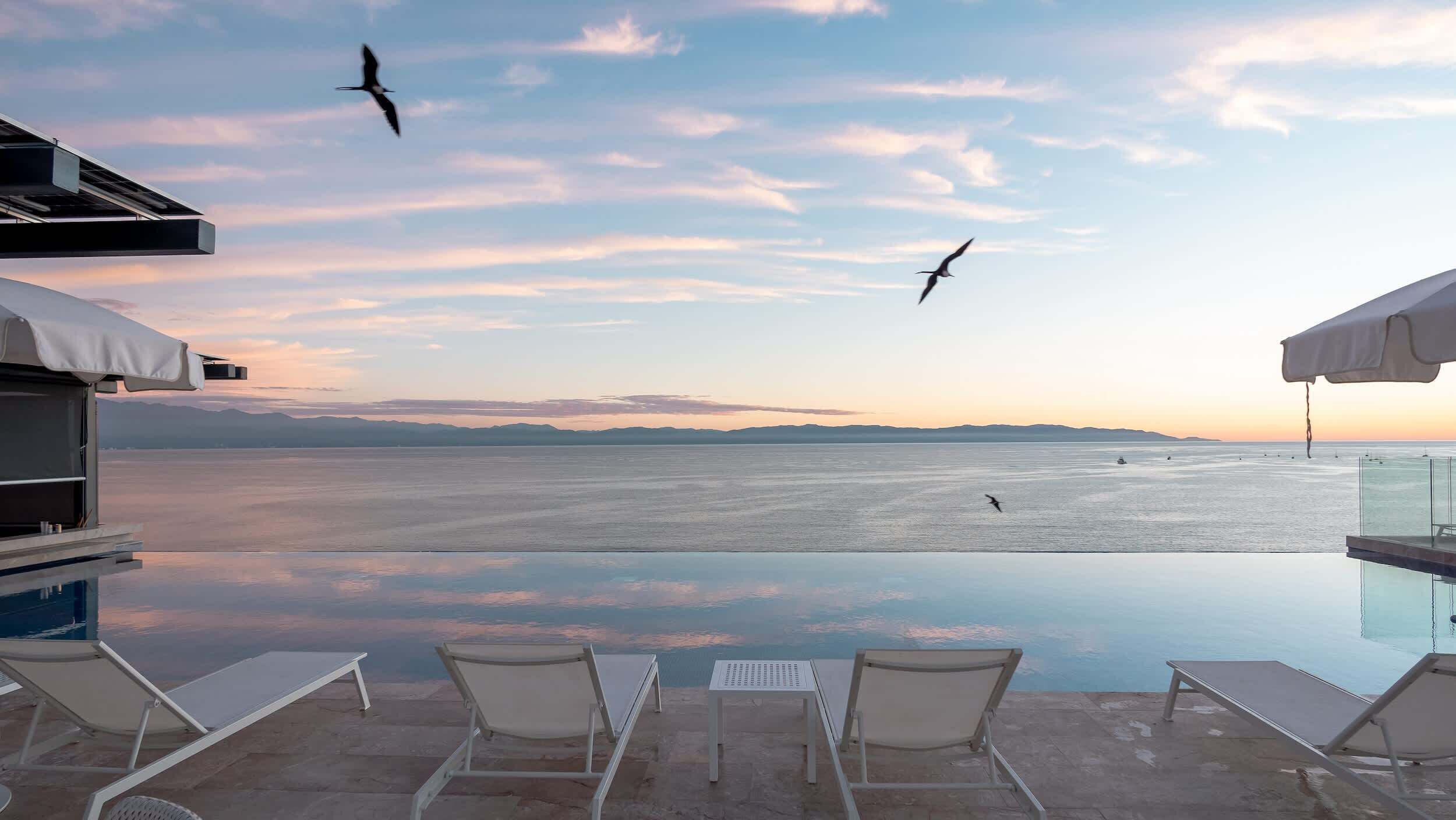 An infinity pool with lounge chairs, a clear view of the sea, and a sunset in the background.