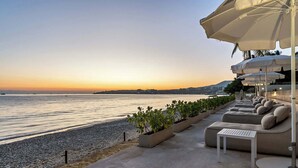 Uma área de estar à beira-mar com mobiliário exterior e guarda-sóis, com vista para um mar tranquilo e um horizonte urbano ao pôr do sol.