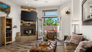 A living room with a fireplace, a television, and a large window with a view of greenery. - Casa Capri-Walking Distance to Old Town LQ + Park (La Quinta)