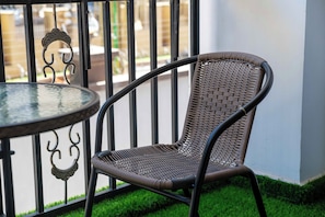 A wicker chair with a curved backrest and a round table with a glass top, positioned on a balcony with a metal railing and a view of a building in the background. - Indulge in luxury: Plush seating, luxurious 7 star mattress. (NAIROBI)