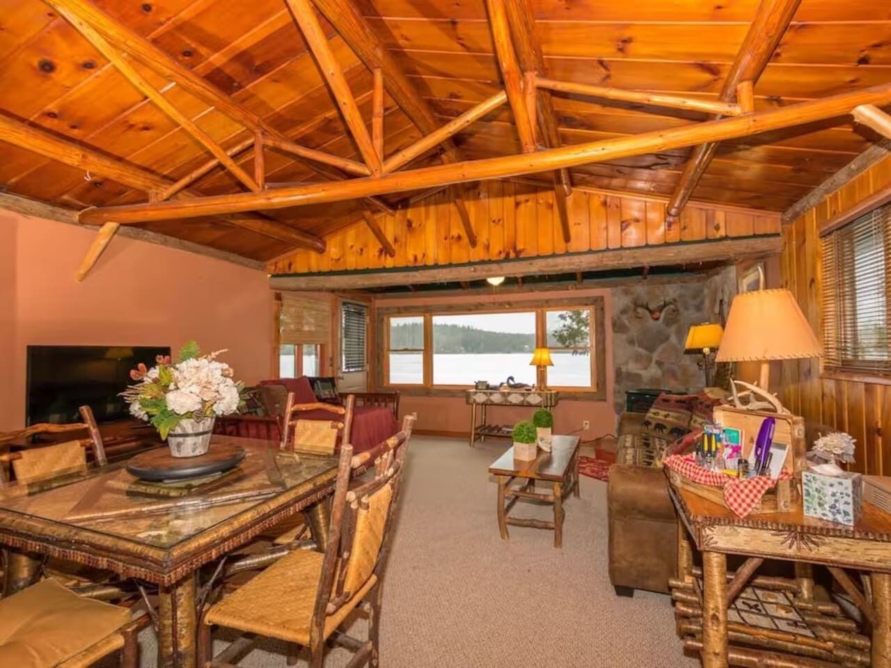 L’intérieur d’un chalet rustique, avec une table à manger en bois, des chaises, un canapé et une vue sur un lac par de grandes fenêtres.