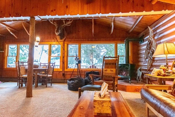 A rustic cabin interior with a wooden ceiling, large windows, a fireplace, and a mounted deer head. - All 4 Cabins on Lake Colby (Saranac Lake)