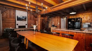 Interior rústico de una cabaña con una mesa de madera para comer, sillas, una araña, una zona de cocina con refrigerador y una chimenea.