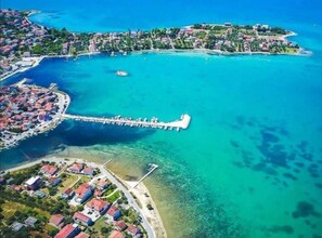 Vista aèria d'una població costanera amb un llarg moll blanc que s'endinsa en un mar turquesa, envoltat d'edificis residencials i aigües blaves cristal·lines.