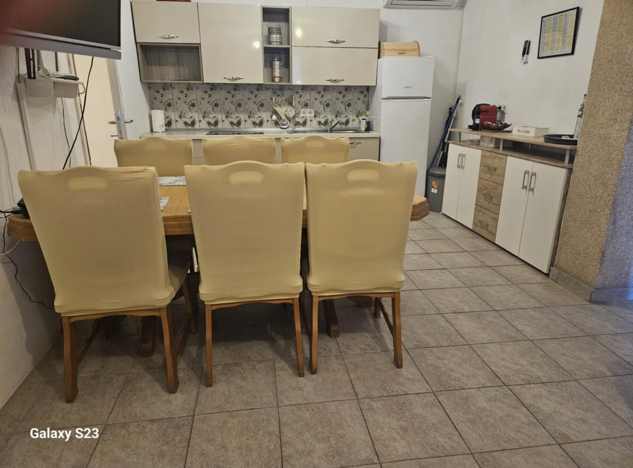 A dining area with a table and six chairs, a kitchen in the background, and a tiled floor.