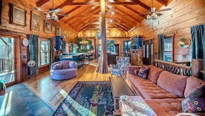 A spacious log cabin interior with a high ceiling, wooden beams, a central seating area with a sofa and armchairs, a dining area, and a kitchen visible in the background. - Vibrant Spacious Mountain Estate with Heated Pool & Hot Tub in Suches (Suches)