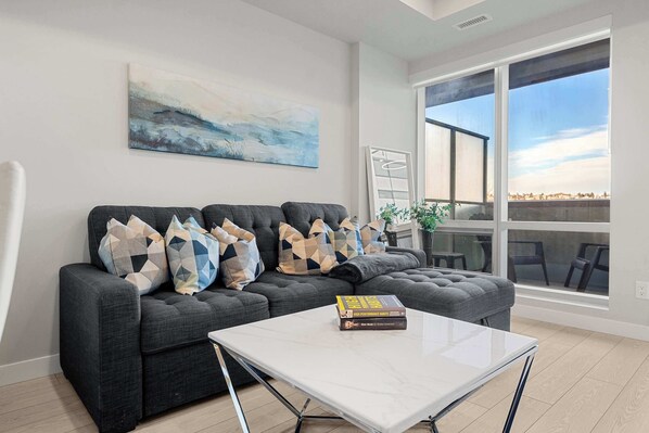 A modern living room with a sectional sofa, a white coffee table, and a large window offering a view.