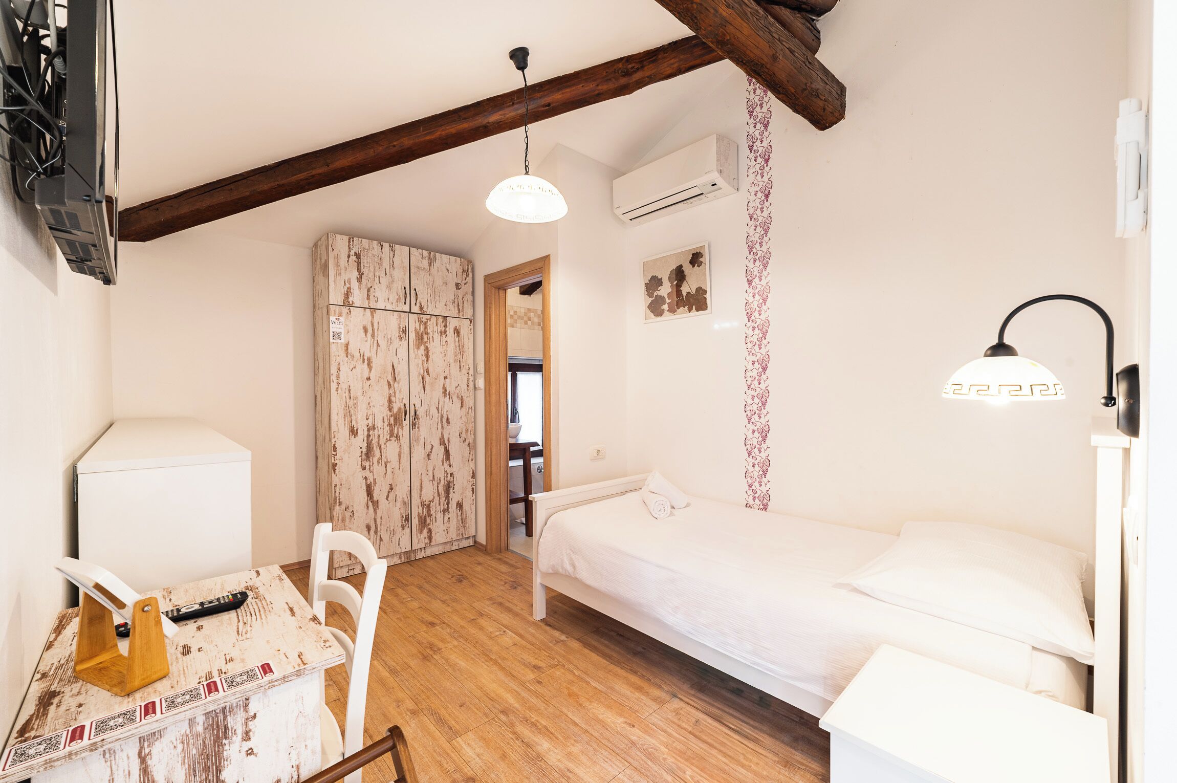 Une petite pièce bien éclairée, avec un lit, un bureau et une chaise. On y trouve une poutre en bois au plafond, un appareil de climatisation et une lampe fixée au mur.
