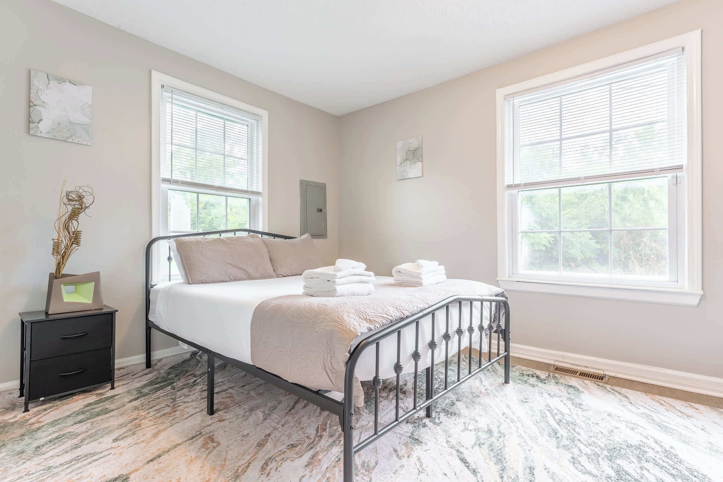 A bedroom with a metal bed frame, a bedside table, a nightstand, and two windows with blinds.
