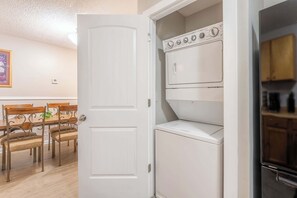 A compact laundry room with stacked washers and dryers, a dining area with a wooden table and metal chairs, and a white door. - The Hope Hideaway - Cozy Home in The Nations (Nashville)