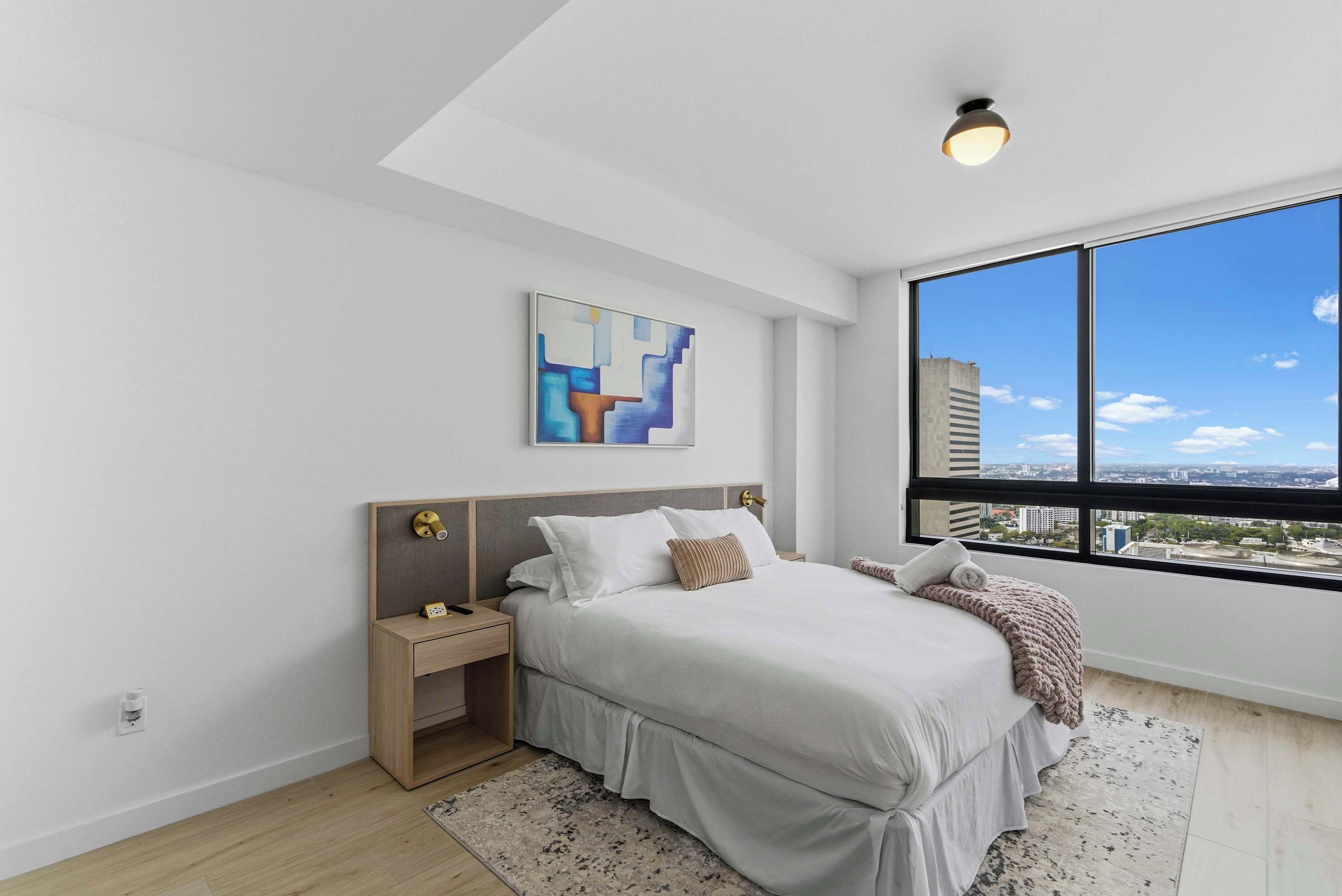 A modern bedroom with a large window, a bed with white bedding, a nightstand, and a wall-mounted abstract art piece.
