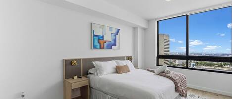 A modern bedroom with a large window, a bed with white bedding, a nightstand, and a wall-mounted abstract art piece.