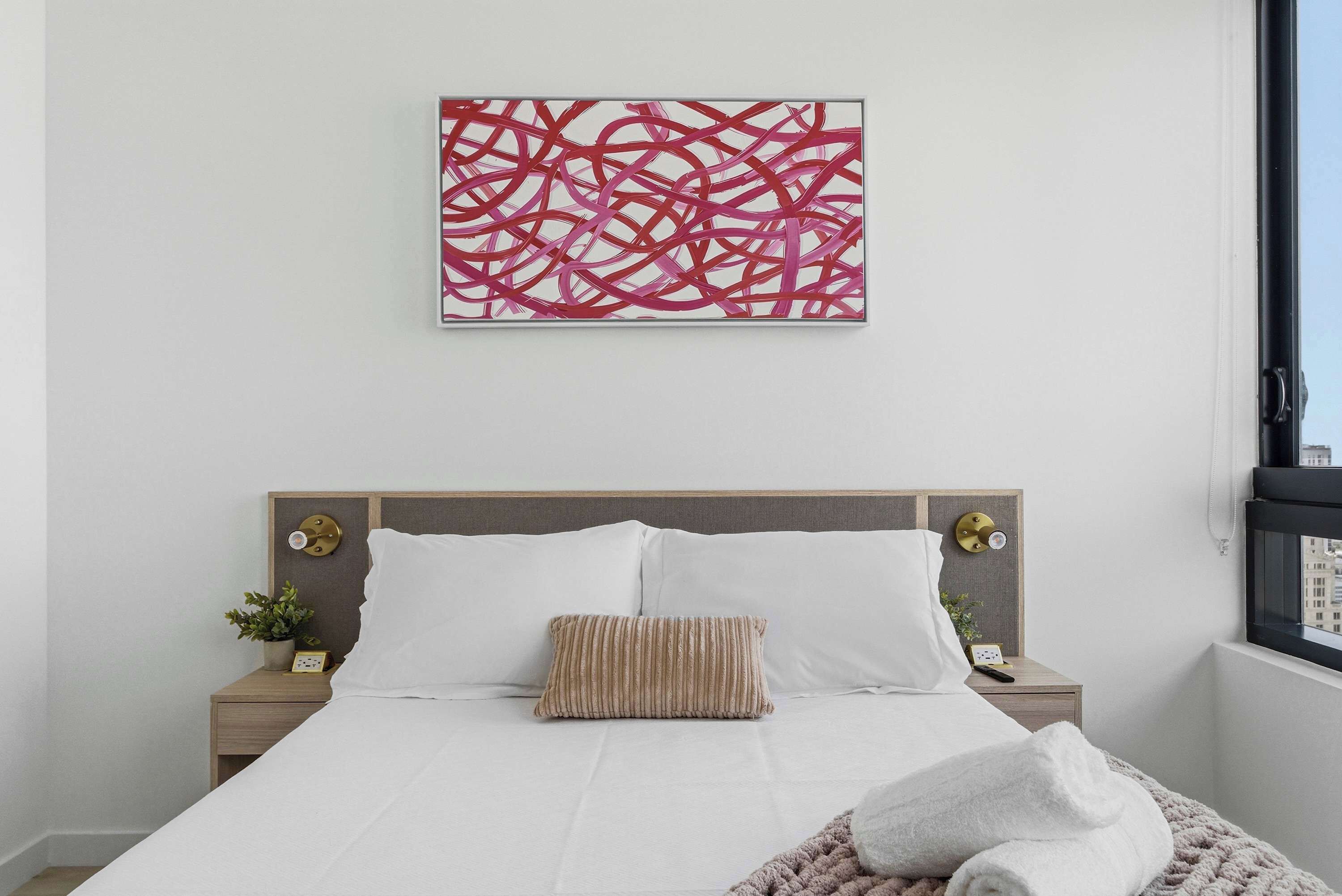 A neatly made bed with white bedding, a striped pillow, and a blanket. There is a wall-mounted abstract art piece above the bed.