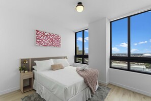 Een moderne slaapkamer met een groot raam, een bed met witte lakens, een nachtkastje met een lamp en een abstract rood schilderij aan de muur.