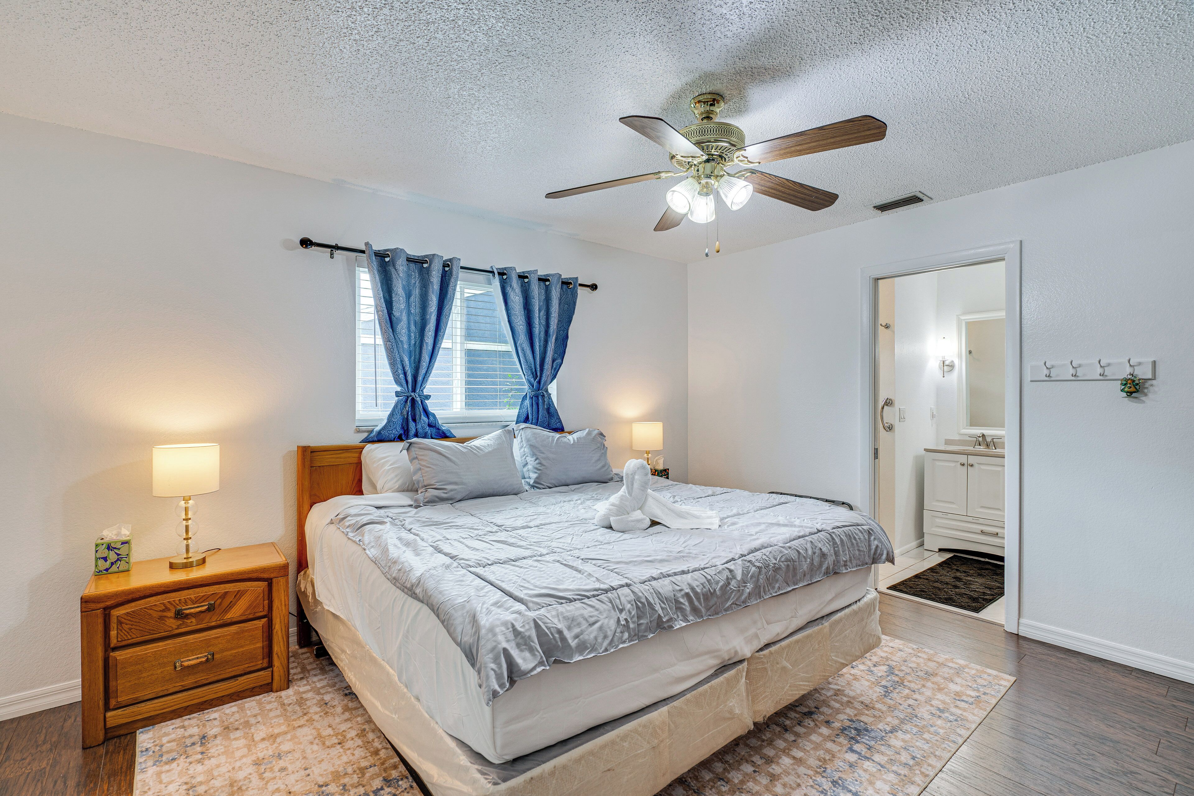 A bedroom with a bed, nightstand, lamp, ceiling fan, and a view of the bathroom.