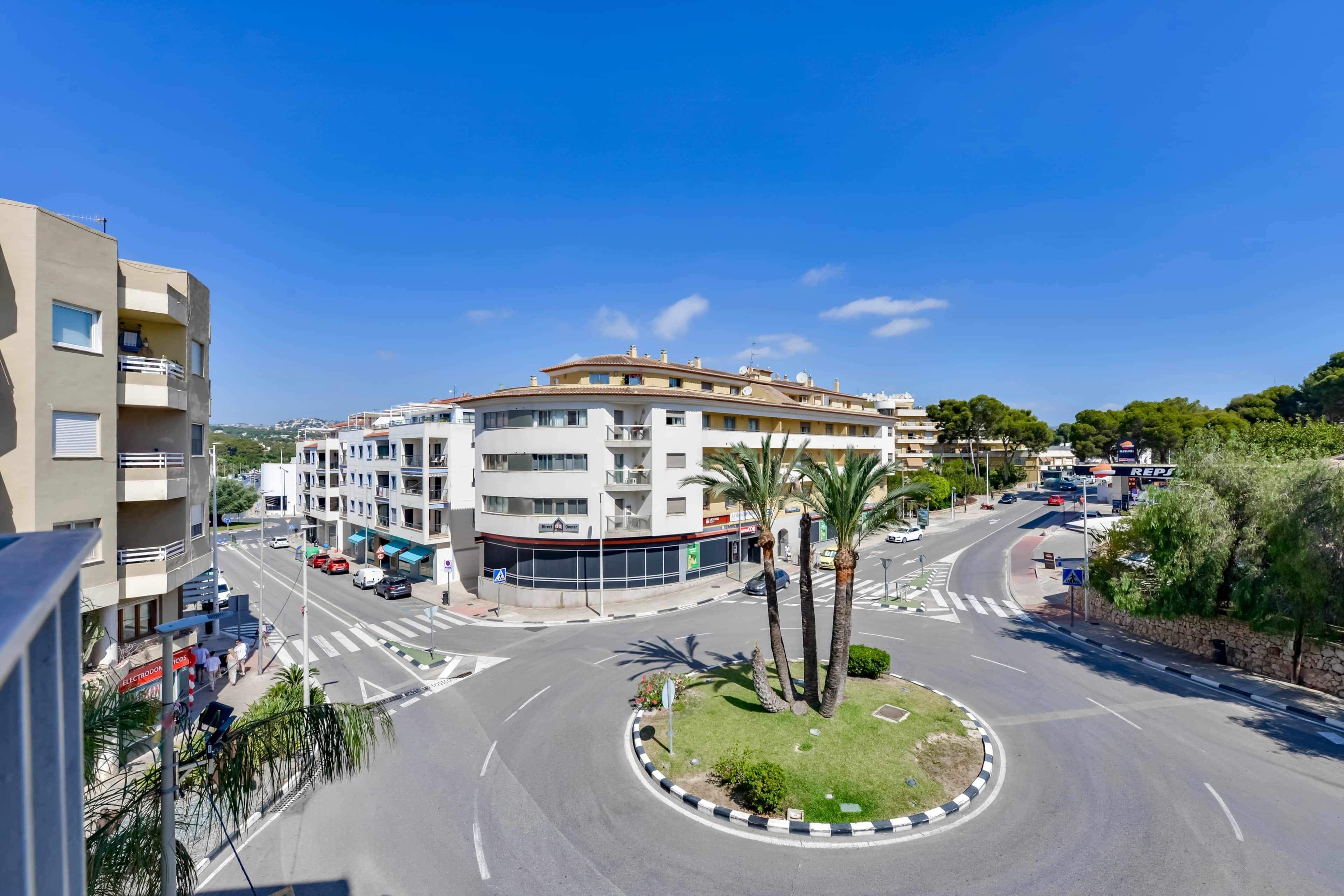 A multi-story building with a curved facade, a roundabout with palm trees, and a clear blue sky.