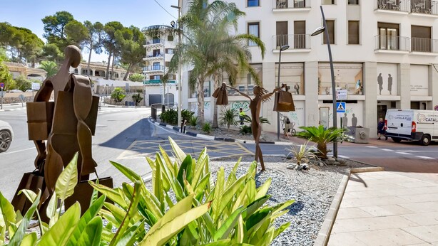 Una calle con una escultura de una figura sosteniendo bolsas, una palmera y un edificio con balcones.