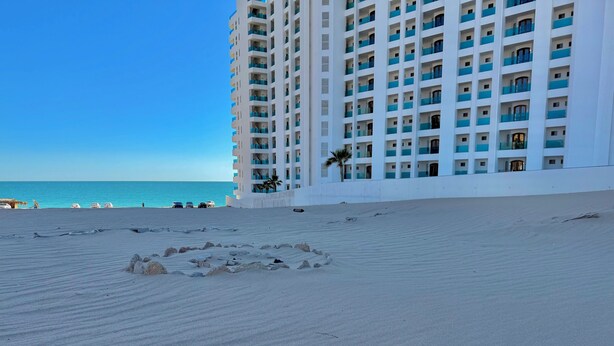 Una playa con un edificio blanco grande, balcones azules, una palmera y un cielo azul despejado.