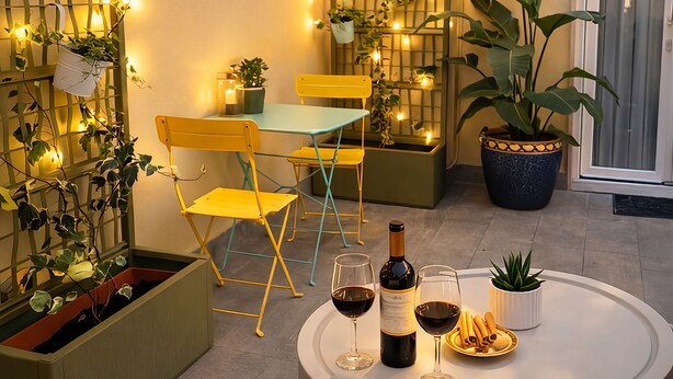 A cozy outdoor seating area with yellow chairs, a table with wine glasses and a bottle, string lights, and potted plants.