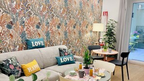 A modern living room with a floral wallpaper, a grey sofa with 'LOVE' and 'HAPPY' cushions, a round dining table with a plant, and a view of the outside.